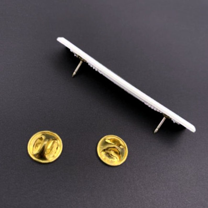 May include: Two gold-colored pin backs, one on the left and one on the right, are shown on a black surface. A white rectangular pin with two gold-colored pin backs is shown above the two pin backs.