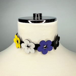 May include: A black choker necklace featuring five colorful flower accents. The flowers are yellow, white, purple, and black, each with a silver center. The necklace is displayed on a mannequin.