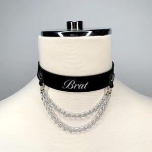 May include: Black leather choker with a silver chain and the word "Brat" engraved on the front.