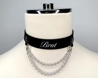May include: Black leather choker with a silver chain and the word "Brat" engraved on the front.