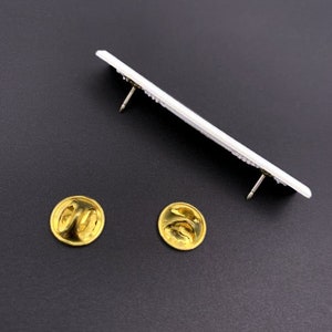 May include: A white metal pin with two gold metal backings. The pin is rectangular and has a raised design.