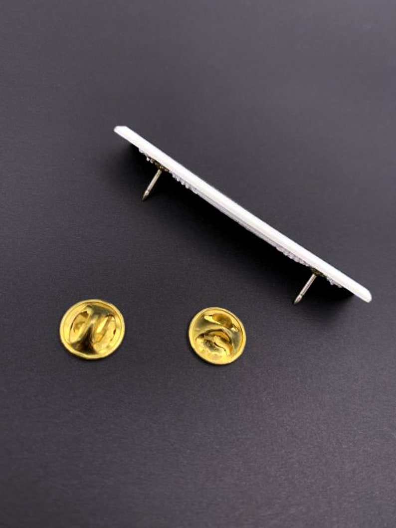 May include: A white metal pin with two gold metal backings. The pin is rectangular and has a raised design.
