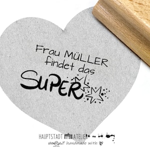 Könnte beinhalten: Ein herzförmiger Stempel mit dem Text "Frau Müller findet das SUPER" in schwarzer Tinte. Der Stempel befindet sich auf einer grauen, strukturierten Oberfläche.
