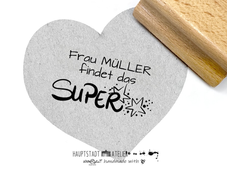 May include: A heart-shaped stamp with the text "Frau M&uuml;ller findet das SUPER" in black ink. The stamp is on a gray textured surface.