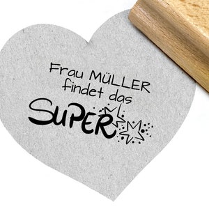 May include: A heart-shaped card with the text "Frau M&uuml;ller findet das SUPER" in black lettering with a star and dots. The card is being stamped with a wooden stamp.