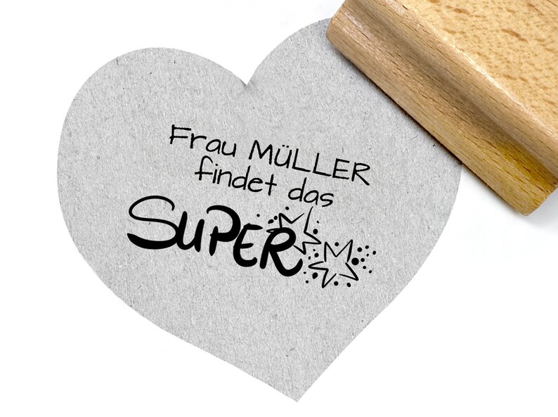 May include: A heart-shaped card with the text "Frau M&uuml;ller findet das SUPER" in black lettering with a star and dots. The card is being stamped with a wooden stamp.