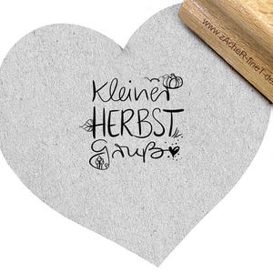 Könnte beinhalten: Ein herzförmiger, grauer Karton mit dem deutschen Schriftzug "Kleiner Herbst Gruß" in schwarzer Schrift, verziert mit kleinen Illustrationen zum Thema Herbst. Ein Holzstempel ist teilweise sichtbar.