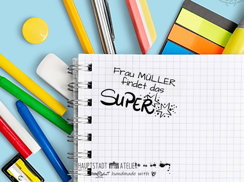 May include: A white notebook with a black spiral binding. The notebook has a black and white graphic of a star with the text "Frau M&uuml;ller findet das SUPER" on the front.