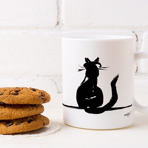 May include: White ceramic mug featuring a black cat silhouette. The cat is depicted from behind. Three chocolate chip cookies are stacked on a white doily beside the mug.