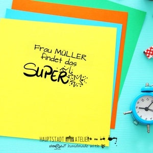 May include: A yellow sticky note with the text "Frau M&Uuml;LLER findet das SUPER" in black lettering with a starburst design. The note is on a blue surface with a blue alarm clock, pencils, and paper clips.