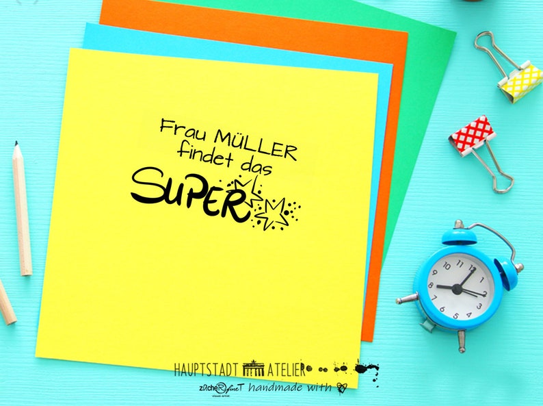 May include: A yellow sticky note with the text "Frau M&Uuml;LLER findet das SUPER" in black lettering with a starburst design. The note is on a blue surface with a blue alarm clock, pencils, and paper clips.