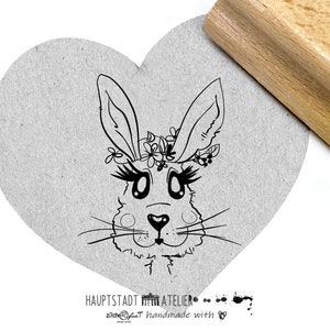 Könnte beinhalten: Schwarz-weiß-Linienzeichnung eines Kaninchenkopfes mit Wimpern und einer Blumenkrone. Das Bild ist auf einem grauen herzförmigen Hintergrund gedruckt. Der Text "HAUPTSTADT ATELIER" und "handmade with ♥" ist unter dem Bild gedruckt.
