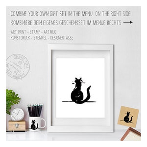 May include: A white framed art print featuring a black cat silhouette, alongside a matching mug and card. The image includes text in English and German, promoting gift sets. The mug and card also display the cat design. The text reads "COMBINE YOUR OWN GIFT SET".