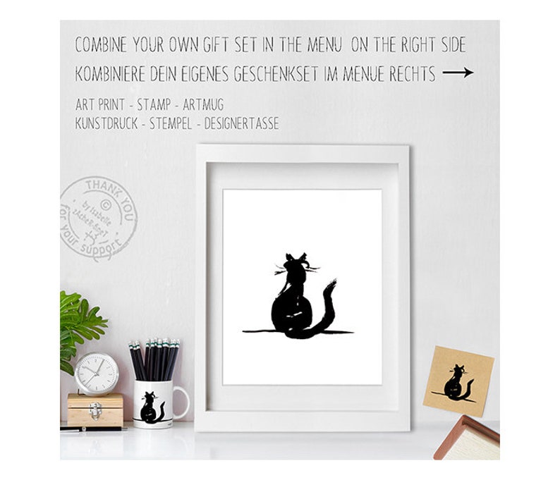 May include: A white framed art print featuring a black cat silhouette, alongside a matching mug and card. The image includes text in English and German, promoting gift sets. The mug and card also display the cat design. The text reads "COMBINE YOUR OWN GIFT SET".