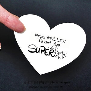 May include: A white heart-shaped card with the text "Frau M&uuml;ller findet das SUPER" in black lettering with a starburst design.