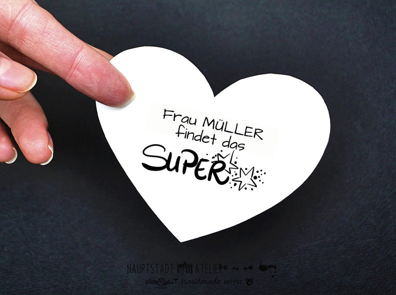 May include: A white heart-shaped card with the text "Frau M&uuml;ller findet das SUPER" in black lettering with a starburst design.