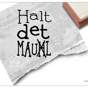 Könnte beinhalten: Ein schwarzer und weißer Stempel mit dem Text "Halt det MAUL" darauf. Der Stempel befindet sich auf einer weißen, strukturierten Oberfläche.