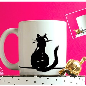 May include: White ceramic mug with a black cat silhouette. The cat is depicted sitting with its tail curled. The mug has a curved handle and is set against a bright pink background.