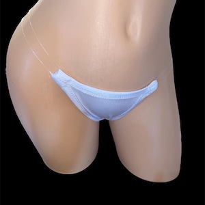 Puede incluir: Tanga blanco con tirantes transparentes. El tanga presenta un pequeño panel frontal triangular y finas tiras transparentes. La prenda se exhibe en un maniquí sobre un fondo negro.