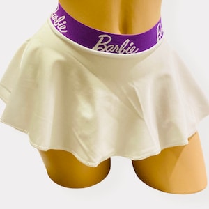 May include: White skirt with a flared design. The waistband is purple with the word "Barbie" in white script. The skirt is displayed on a mannequin.