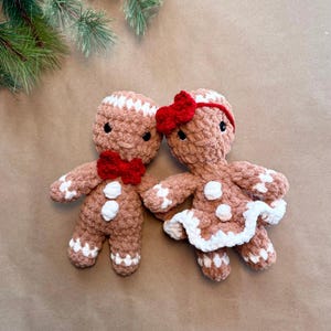 May include: Two crocheted gingerbread dolls made of brown yarn with white accents. One doll wears a red bow tie, while the other features a red hair bow and a white ruffled skirt. Both dolls have black eyes and white frosting-like details.