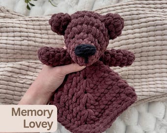 May include: A dark brown crochet bear lovey made of soft, chunky yarn. The bear features a rounded head with small ears, short arms, and a black stitched nose. The body consists of a textured, square-shaped blanket base. Text reads Memory Lovey.