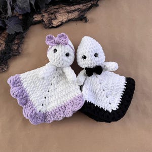 May include: Two crochet ghost loveys made of white yarn. The left ghost features a light purple bow and a matching light purple ruffled border. The right ghost wears a black bow tie and has a black ruffled border. Both have black safety eyes.