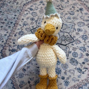 May include: A handmade crochet duck doll with a white body, yellow beak, and matching boots. The duck wears a green party hat with a white pom-pom and a yellow ruffled collar. The doll is soft and plush.