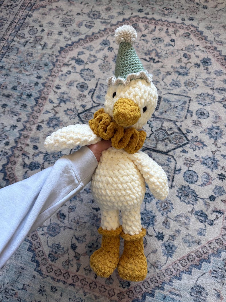 May include: A handmade crochet duck doll with a white body, yellow beak, and matching boots. The duck wears a green party hat with a white pom-pom and a yellow ruffled collar. The doll is soft and plush.