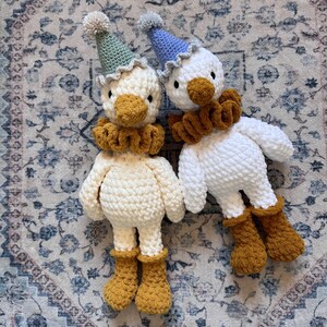 May include: Two handmade crochet duck dolls. One is cream-colored, the other is white. Both have yellow-orange beaks, ruffs, and boots. They wear party hats with pom-poms. The dolls are lying on a patterned rug.