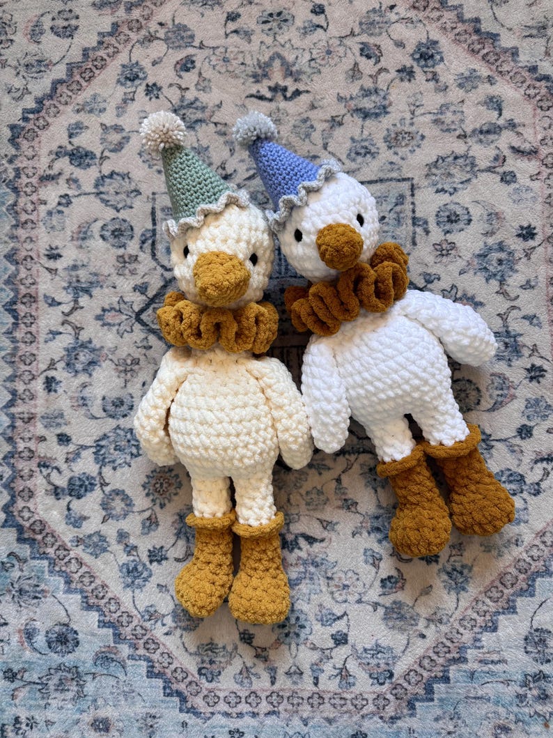 May include: Two handmade crochet duck dolls. One is cream-colored, the other is white. Both have yellow-orange beaks, ruffs, and boots. They wear party hats with pom-poms. The dolls are lying on a patterned rug.