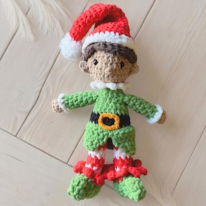 Può includere: Una bambola elfo all'uncinetto con un cappello rosso e bianco con pompon, capelli castani e viso beige. L'elfo indossa una tunica verde con cintura nera e fibbia gialla, un colletto bianco e leggings a righe rosse e bianche con scarpe verdi.