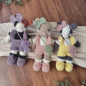 May include: Three handmade crochet plush animals wearing outfits and bows. The left one is black and white with purple overalls and shoes. The middle one is tan with pink overalls and a green bow. The right one is gray and white with yellow overalls.