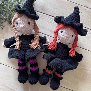 May include: Two handmade crochet witch dolls. Each doll has a black witch hat, black dress, and braided hair. One doll has tan braids, the other has red braids. The dolls have black shoes and striped socks in purple and orange.