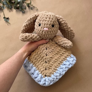 May include: A crochet security blanket featuring a tan plush bunny head with long ears, black safety eyes, and a small pink stitched nose. The attached blanket body is made of chunky tan yarn with a light blue border along the bottom edge.