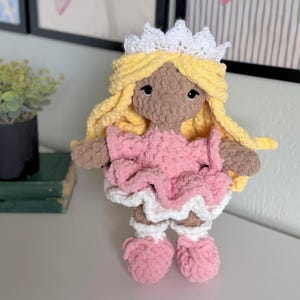 May include: A handmade crochet doll with brown skin, long yellow yarn hair, and a white crown. The doll wears a pink dress with a ruffled white hem and matching pink shoes with white cuffs. The doll has simple black embroidered eyes and a soft texture.