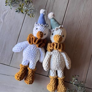 May include: Two handmade white crochet duck dolls with orange beaks, ruffs, and boots. Each duck wears a party hat with a pom-pom. One hat is blue, the other is green. The dolls are on a light wood surface.