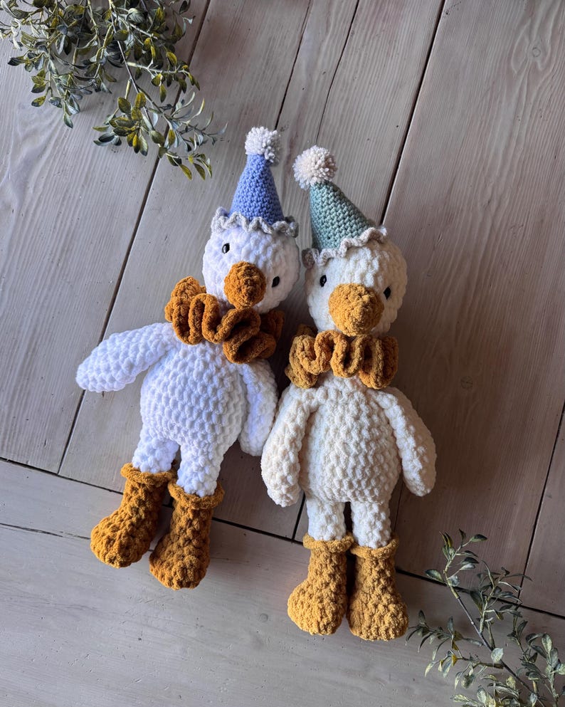 May include: Two handmade white crochet duck dolls with orange beaks, ruffs, and boots. Each duck wears a party hat with a pom-pom. One hat is blue, the other is green. The dolls are on a light wood surface.