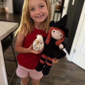 Young girl holding a handmade crochet witch doll and plush chick, showing the finished size of the Halloween crochet pattern.
