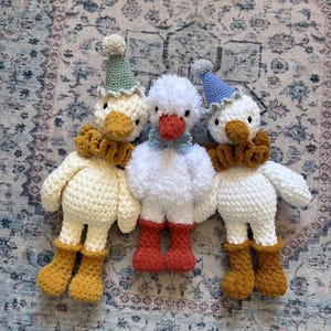 May include: Three handmade crochet duck toys. The ducks have white bodies, orange beaks, and are wearing hats and ruffs. The duck on the left has a green hat and mustard-colored boots. The center duck has red boots. The duck on the right has a blue hat and mustard-colored boots.