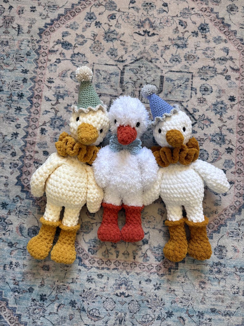 May include: Three handmade crochet duck toys. The ducks have white bodies, orange beaks, and are wearing hats and ruffs. The duck on the left has a green hat and mustard-colored boots. The center duck has red boots. The duck on the right has a blue hat and mustard-colored boots.