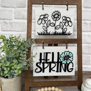 Puede incluir: Un letrero decorativo de madera con dos paneles blancos. El panel superior presenta recortes de flores negras sobre hierba. El panel inferior dice "HELLO SPRING" con un acento de flor turquesa. El letrero es marrón y tiene un marco en forma de escalera.