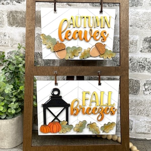 May include: A wooden easel with two signs. The top sign says "autumn leaves" with a fall-themed design. The bottom sign says "fall breezes" with a lantern and pumpkins.