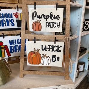 May include: A wooden easel with two signs. The top sign says "Pumpkin Patch" with a pumpkin graphic. The bottom sign says "U-Pick 5¢" with two pumpkins, one orange and one white.