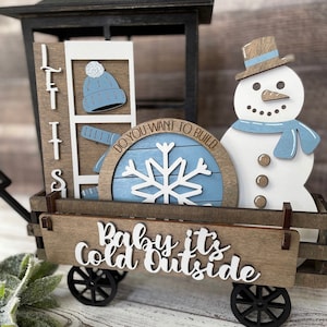 Puede incluir: Un carro de madera con un muñeco de nieve, un copo de nieve y un sombrero en la parte superior. El carro tiene un letrero que dice "Baby it's Cold Outside".
