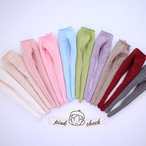 Könnte beinhalten: Eine Auswahl an Puppen-Leggings in verschiedenen Farben: Weiß, Rosa, Blau, Grün, Lila, Beige, Rot und Grau. Jedes Paar hat ein Zopfmuster. Das Logo "pink cheek" ist sichtbar.