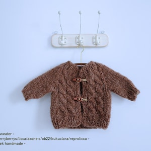 May include: A brown knitted sweater with a cable knit design and wooden toggle closures. The sweater is hanging on a hanger against a white wall. The text "- brown sweater -" is visible.