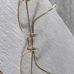 May include: Gold-rimmed eyeglasses with clear lenses. The frames have a delicate, vintage design with a bridge connecting the two lenses. The temples are a light color and curve gently to fit over the ears. The glasses are resting on a white surface.