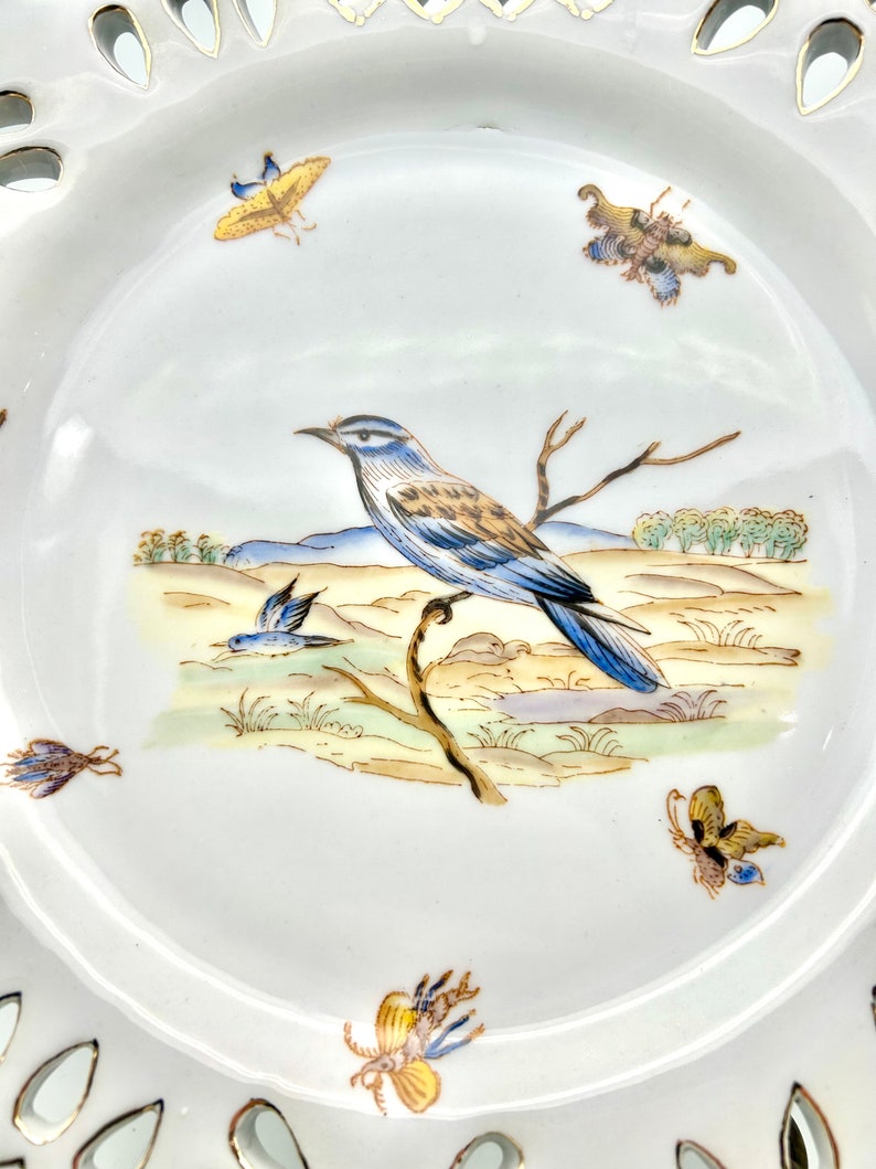 May include: White porcelain plate with a detailed bird and butterfly design. The plate features a central image of a blue and brown bird perched on a branch, surrounded by painted butterflies. The rim has a decorative, openwork design.