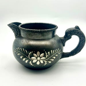 Könnte beinhalten: Antike silberfarbene Sahnekännchen mit floralem Design. Das Kännchen hat einen Ausguss, einen dekorativen Griff und einen geriffelten Rand. Das weiße Blumendesign ist auf der Vorderseite zentriert. Das Kännchen steht vor weißem Hintergrund.